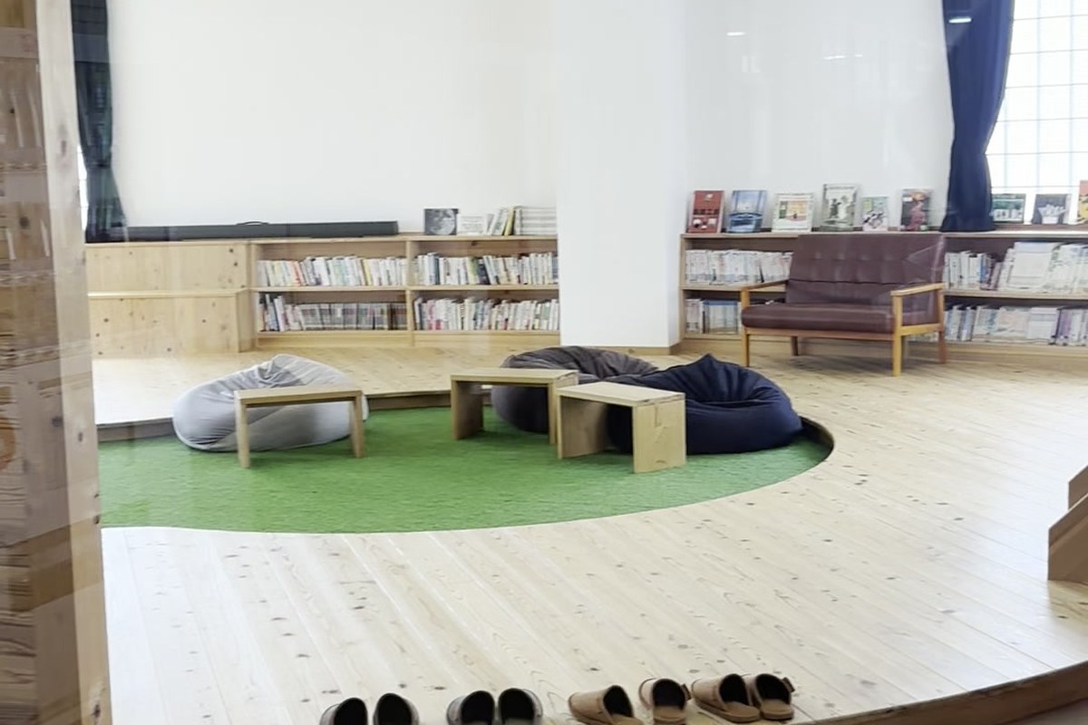 FOREST DOOR 旧神楽小学校の木の床のくつろぎスペース。丸い木のステージに人工芝とビーズクッション、壁際の本棚