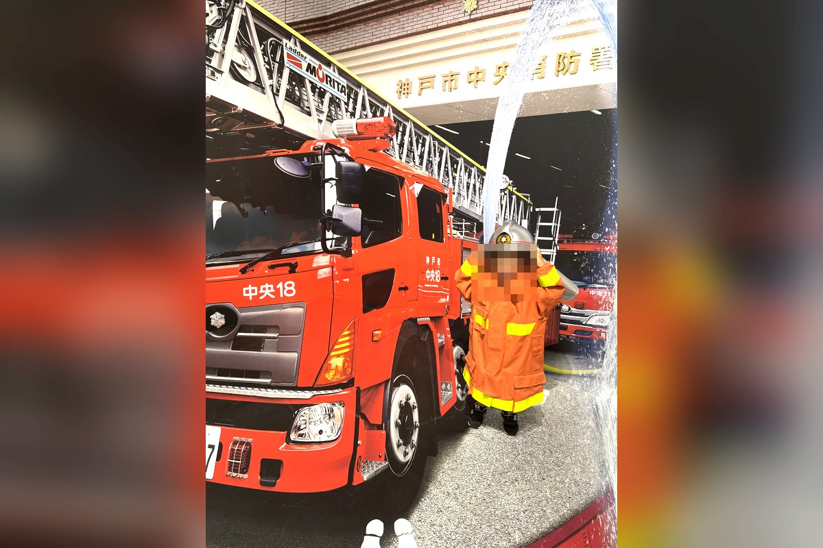 バンドー神戸青少年科学館で、子ども用の消防服とヘルメットを着て消防車の写真パネルの前に立つ3歳の子ども
