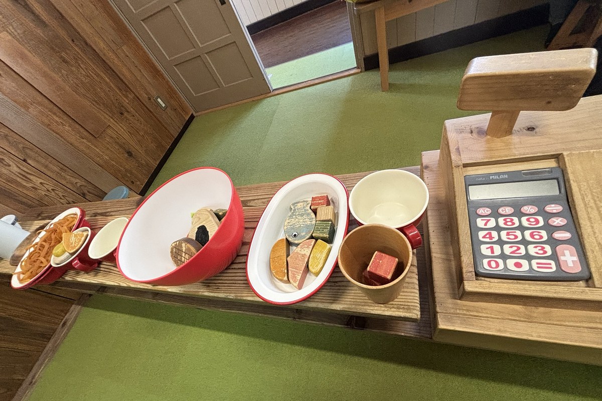 おままごとカフェのカウンター。パスタやお魚の木のおもちゃが入ったお皿と、電卓つきの木のレジ