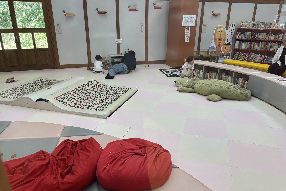 絵本の部屋の全景。マットの床に大きな絵本とワニのクッション、壁ぎわに絵本の棚