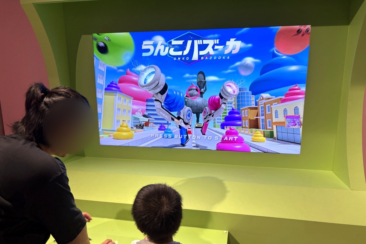 クソゲーセンターの「うんこバズーカ」の画面の前で、ボタンに手を置いてゲームを始める4歳の後ろ姿と、横で見守る父親